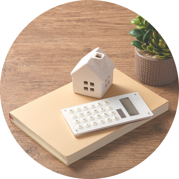木製テーブルの上に置かれた本と電卓、小さな家の模型と観葉植物が並ぶ住宅購入や資金計画のイメージ